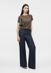 Žena u smeđoj majici kratkih rukava, tamnim hlačama širokih nogavica, crnim sandalama s otvorenim prstima i zelenom torbom na ramenu, stoji na jednobojnoj bijeloj pozadini.