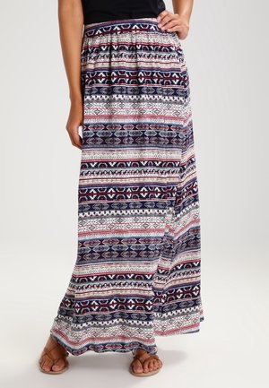 Femme portant une longue jupe à motifs avec des rayures géométriques bleu marine, blanches et rouges, associée à des sandales beiges et un haut noir.