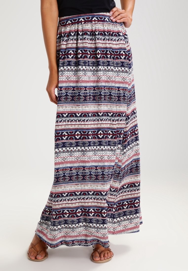 Femme portant une longue jupe à motifs avec des rayures géométriques bleu marine, blanches et rouges, associée à des sandales beiges et un haut noir.