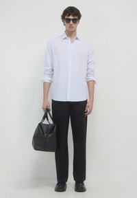 Homme portant des lunettes de soleil, une chemise à rayures blanches à manches longues, un pantalon noir et des chaussures noires, tenant un grand sac en cuir noir.