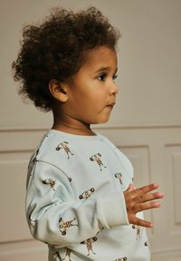 Kääbus, kel on lokkis juuksed, seljas helesinine sviiter, millel on koomiksikujulised jõutõstjad, seisab paremale poole, käsi üles tõstetud neutraalse paneelitud taustaga.