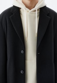 Blazer in misto lana nera con rever a punta, due bottoni e finitura testurizzata, indossato sopra un hoodie beige con lacci.