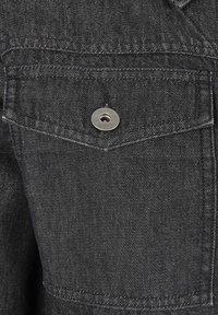 Svart denimtyg med en texturerad yta. Har ett bröstfack med en silverfärgad metallknapp och kontrasterande sömdetaljer.