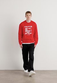 Sarkana džemperis ar baltu tekstu un grafisko dizainu, kurā redzams vārds "CALVIN" un horizontālas līnijas. Pāris ar melnām biksēm un baltām sporta apaviem.