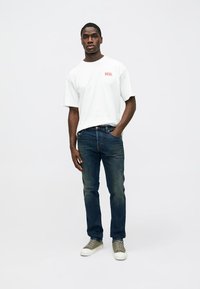 Camiseta blanca de algodón con el logo "DIESEL" en rojo, combinada con jeans azul oscuro y zapatillas grises. El modelo está de pie con las manos en los bolsillos.