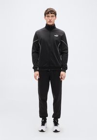 Μαύρο αθλητικό μπουφάν με ψηλό γιακά, κλείσιμο με φερμουάρ και λευκές ραφές, σε συνδυασμό με μαύρο παντελόνι και μαύρα-λευκά αθλητικά παπούτσια.