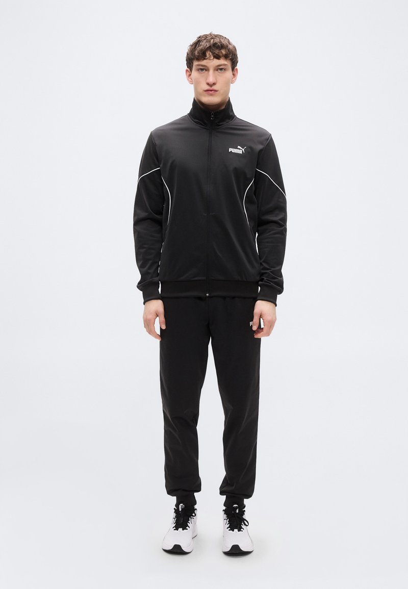 Μαύρο αθλητικό μπουφάν με ψηλό γιακά, κλείσιμο με φερμουάρ και λευκές ραφές, σε συνδυασμό με μαύρο παντελόνι και μαύρα-λευκά αθλητικά παπούτσια.