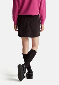 Černá džínsová minisukně s jednoduchým designem, spárovaná s černými podkolenkami a sandály. Pozadí je jednoduše bílé.