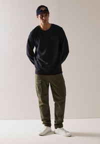 Granatowa koszula z długim rękawem, oliwkowe spodnie cargo z kieszeniami, białe sneakersy oraz granatowa czapka z czerwonymi akcentami. Prosty i funkcjonalny design.