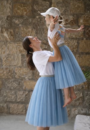 Frau hebt lächelndes Mädchen mit weißer Kappe, beide in weißen Oberteilen und passenden blauen Tüllröcken, vor einem steinernen Wandhintergrund.
