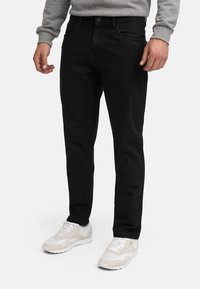 Mann trägt schwarze Slim-Fit-Jeans, grauen Pullover und weiße Turnschuhe, steht vor einem einfarbigen Hintergrund.