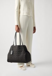 Black quilted tote bag with double handles, featuring floral pattern and a small tag. Structured shape with soft texture.