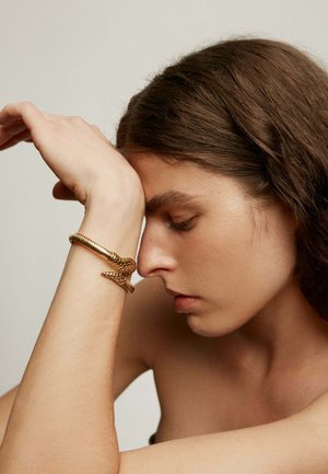 Mujer con los ojos cerrados apoyando la frente en el brazo, llevando una pulsera dorada con forma de serpiente sobre un fondo liso.