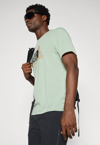 Camiseta de algodón verde claro con un logotipo gráfico en negro y naranja, llevada con una mochila negra. El modelo también lleva gafas de sol oscuras.