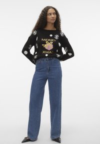 Svart genser med hvite snøfnugg og prikker, med teksten "MERRY XMAS" i gull og et rosa gave-design. Kombinert med høytlivede blå jeans.