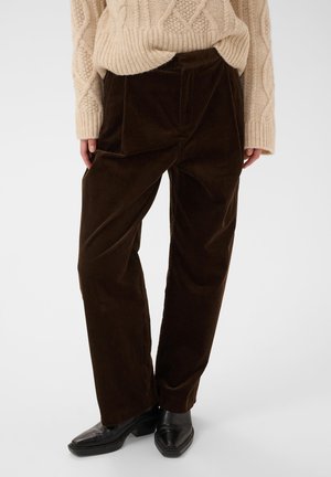 Personne portant un pantalon en velours côtelé marron foncé, un pull à torsades beige et des chaussures en cuir noir, debout sur un fond uni.