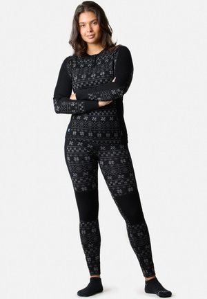Mujer de pie con los brazos cruzados, vestida con camiseta térmica negra y leggings con patrones de copos de nieve escandinavos grises.