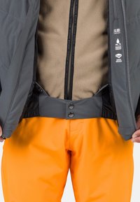Grau wasserdichte Jacke, die über ein beige Fleece-Futter geöffnet ist, kombiniert mit knallorangefarbenen Hosen. Verfügt über einen Druckknopfverschluss und winddichte Merkmale.