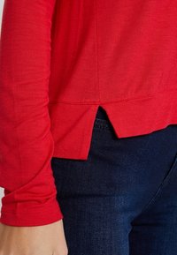 Rotes Langarmoberteil mit strukturierter Oberfläche, das einen asymmetrischen Saum und seitliche Schlitzdetails aufweist. Kombiniert mit dunkelblauen Jeans.