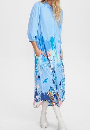 Frau trägt ein hellblaues, langärmeliges Midikleid mit einem mehrfarbigen abstrakten Meeresmuster und weißen kniehohen Stiefeln, die Hände in den Taschen.