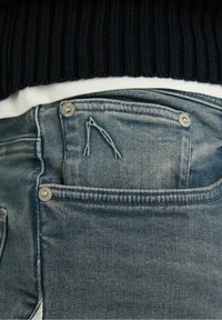 Jeans di denim azzurro chiaro con design standard delle tasche, bottoni in metallo argentato e un motivo di cucitura distintivo vicino alla tasca.