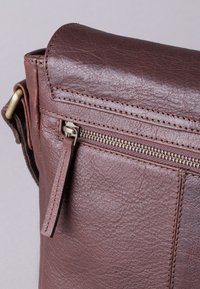 Brown leather bag with a textured finish, featuring a zippered pocket and a top flap. Brass zipper hardware and stitching details are visible.