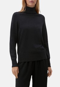 Pull à col roulé noir en tissu doux, avec manches longues et coupe décontractée. Porté avec un pantalon noir texturé.