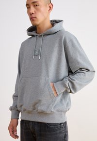 Sudadera con capucha gris de tejido suave, con un bolsillo frontal, que presenta un parche con el logo y cuerdas ajustables con puntas metálicas.