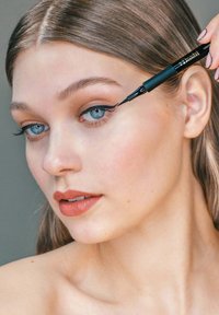 Zwarte eyelinerpen wordt aangebracht op het bovenste ooglid van een persoon met een lichte huid, blauwe ogen en nude lippenstift. Glanzend haar.