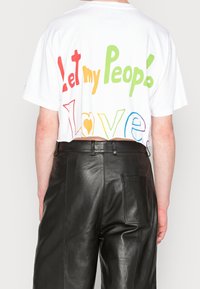 Vit bomullst-shirt med kort ärm som har flerfärgad text på baksidan som säger "Låt mitt folk älska" med varierande typsnitt. Svarta läderbyxor.