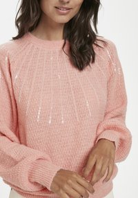 Frau trägt einen weichen, rosa gestrickten Pullover mit subtilen vertikalen Pailletten, langen Ärmeln und umgeschlagenen Handgelenken, die Hände behutsam gefaltet.