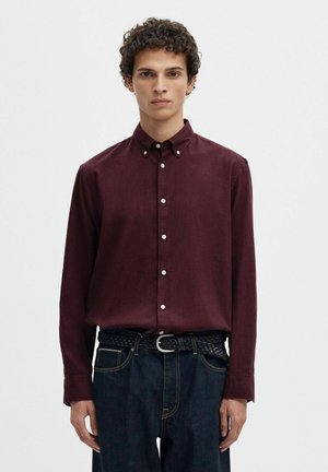 Jeune homme aux cheveux courts et bouclés portant une chemise bordeaux à manches longues boutonnée, rentrée dans un jean foncé avec une ceinture noire tissée.