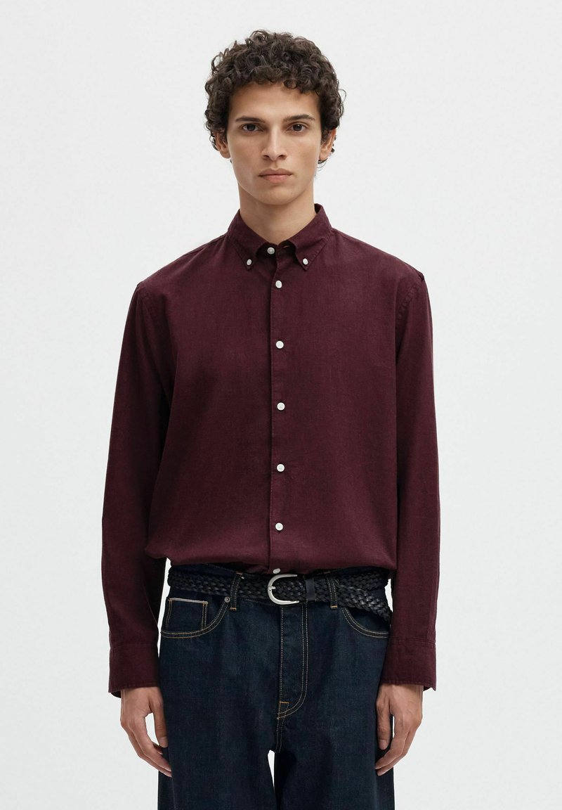Jeune homme aux cheveux courts et bouclés portant une chemise bordeaux à manches longues boutonnée, rentrée dans un jean foncé avec une ceinture noire tissée.