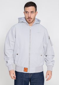 Hellgraue Bomberjacke mit verstellbarer Kapuze, Reißverschluss vorne, seitlichen Taschen und gerippten Bündchen. Verfügt über ein Logo auf der Brust und ein Etikett am Saum.