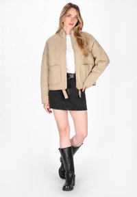 Beige gesteppte Jacke mit Stehkragen, Fronttaschen und cropped Design, kombiniert mit einem schwarzen Jeansrock und schwarzen knielangen Stiefeln.