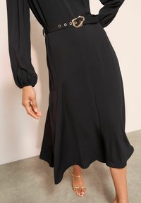 Čern�é šaty s dlouhými rukávy, hladkým povrchem, zúženým pasem a zlatým páskem s přezkou. Volně splývavý A-line lem s jemným drapériem. Nosené se zlatými sandály.