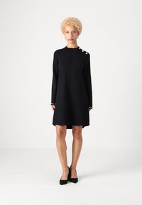 Čierne šaty s dlhými rukávmi a rovnými líniami, zdobené dekoračnými gombíkmi na jednom pleci a s zaobleným lemom. Hladká textúra.