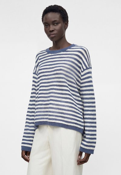 Γυναίκα που φοράει άνετο μπλε και λευκό πουλόβερ με οριζόντιες ρίγες και μακριά μανίκια και λευκό παντελόνι με φαρδιά πόδια, στέκεται μπροστά σε απλό φόντο.