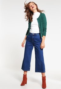 Grønn strikket cardigan med knappedetaljer, hvit høyhalset skjorte, høy midje blå denim culottes, og rustfargede ankelstøvler.