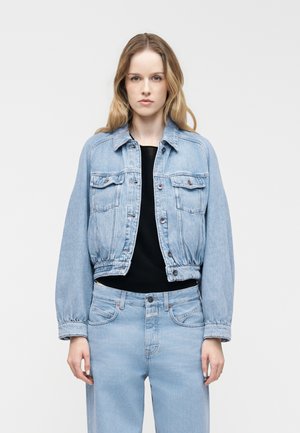 Kobieta ubrana w jasnoniebieską kurtkę dżinsową i dopasowane jeansy, pod którą ma czarną koszulę, stoi na tle jasnego, jednolitego tła.