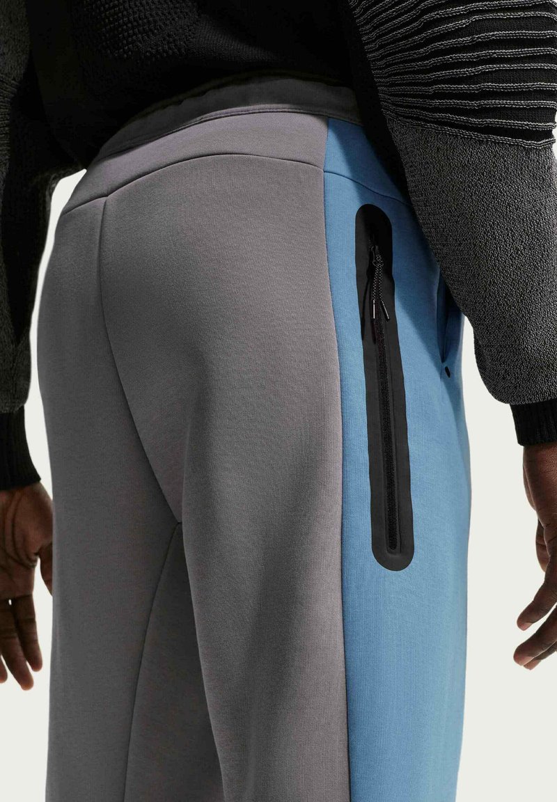 Nahaufnahme einer Person, die zweifarbige graue und blaue Hosen mit einer vertikalen schwarzen Reißverschlusstasche an der Seite und einen schwarzen strukturierten Pullover trägt.