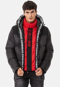 Schwarze Pufferjacke mit rotem Innenfutter, ausgestattet mit einem hohen Kragen, auffälligen Logopunkten und einem Reißverschluss vorne mit einem Farbblockdesign.