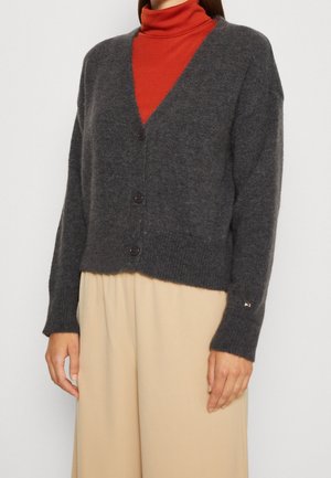 Cardigan en laine gris charbon avec un col en V, des boutons à l'avant, des poignets et un ourlet côtelés. Associé à un pull à col roulé rouge et un pantalon large beige.