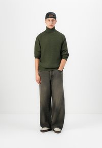 Junger Mann, der eine schwarze rückwärts getragene Kappe, einen grünen Rollkragenpullover, weit geschnittene ausgebleichte schwarze Jeans und weiße Turnschuhe trägt, steht mit einer Hand in der Tasche.