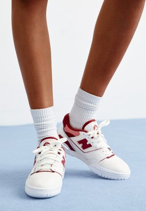 Baskets blanches avec des accents rouges, présentant une tige en cuir perforé, une fermeture à lacets et une semelle en caoutchouc texturée, associées à des chaussettes blanches montantes.