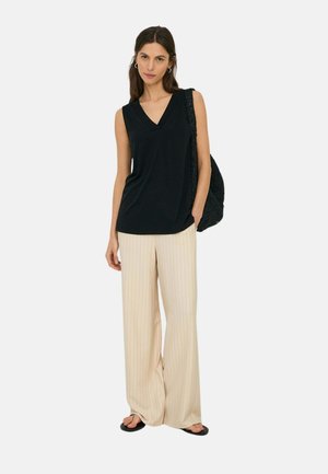 Mujer con top negro sin mangas y escote en V, pantalones beige de pierna ancha con rayas finas, sandalias negras, llevando un bolso de hombro negro con textura.