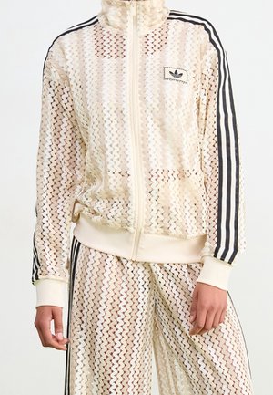Personne portant une veste de survêtement beige en dentelle avec fermeture éclair intégrale et un pantalon assorti avec les trois bandes noires Adidas aux manches et aux jambes.