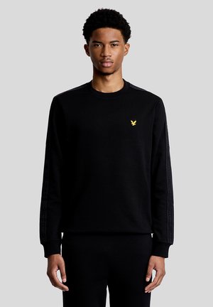 Junger Mann, der einen schwarzen Pullover mit einem kleinen gelben Vogel-Logo auf der Brust und markierten Streifen an den Ärmeln trägt.