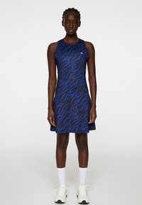 Niebieska i czarna sukienka w wzór tygrysich pasków, bez rękawów, delikatnie rozszerzana u dołu, z logo. Noszona z białymi skarpetkami i trampkami.