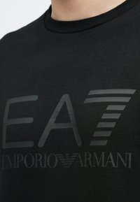 Mustast puuvillast t-särk, millel on suur, tekstuuriga tumehall EA7 logo ja rinnal sujuva fontiga "Emporio Armani" tekst.